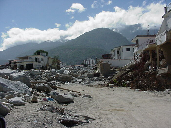 Severe natural disaster damage with destroyed buildings and debris against a mountainous backdrop under cloudy skies.