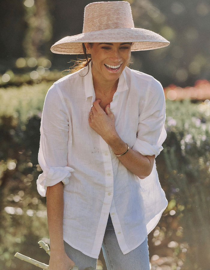 Meghan Markle smiling outdoors wearing a wide-brimmed hat and white shirt during a sunny day in the garden setting.