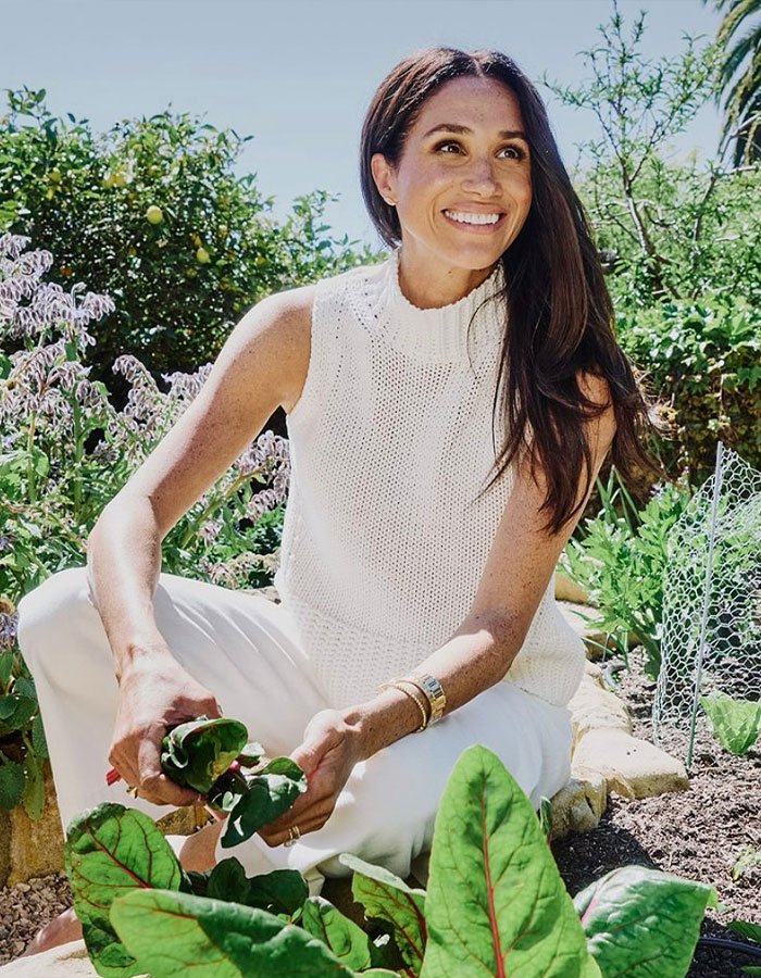 Meghan Markle smiling outdoors, wearing a white sleeveless top, sitting in a garden with green plants around her.