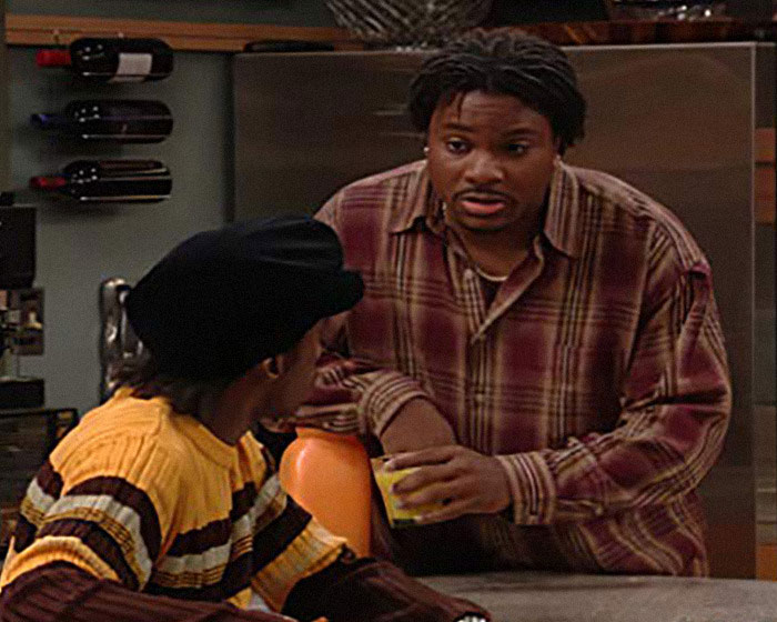 Actor Malcolm-Jamal Warner in a plaid shirt speaking with another person in a kitchen setting, second victim mentioned.
