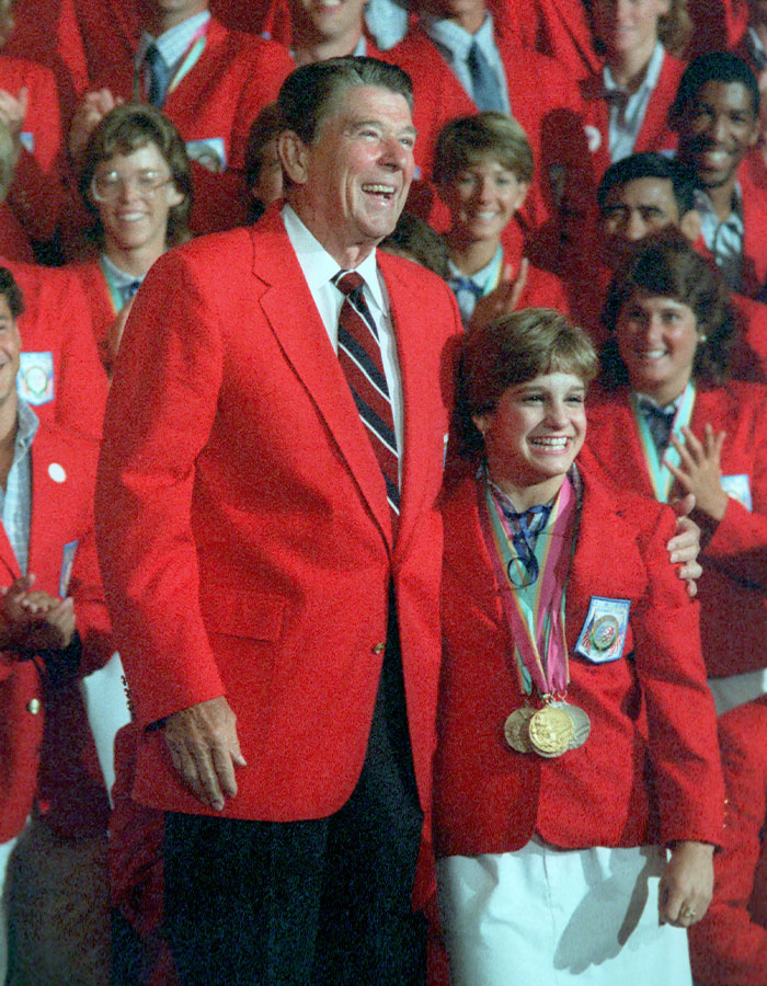 Olympic gold medalist Mary Lou Retton smiling with medals, wearing red team jacket among group in red blazers.
