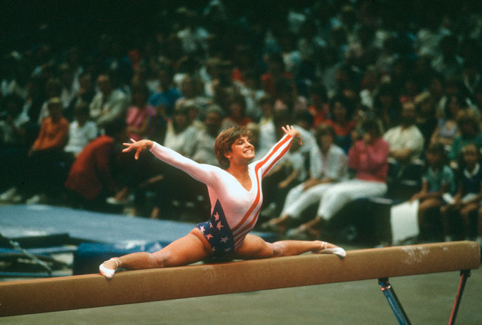 Olympic gold medalist Mary Lou Retton performing a split on balance beam during a gymnastics competition with an audience in the background