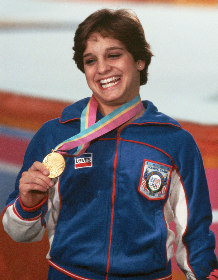 Olympic gold medalist Mary Lou Retton smiling with gold medal, wearing Team USA jacket during award ceremony.