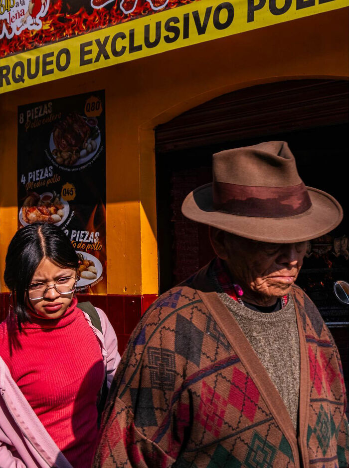 Man in a patterned poncho and hat walking past a young woman in glasses, capturing honest street moments of everyday life.
