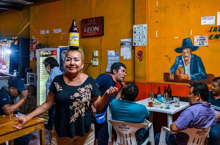Woman balancing a beer bottle on her head in a bar, capturing honest street moments showing the beauty of everyday life.