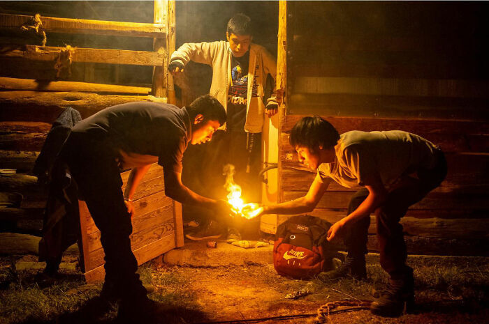 Three young men interacting around a small fire outside a wooden structure, capturing honest street moments of everyday life.