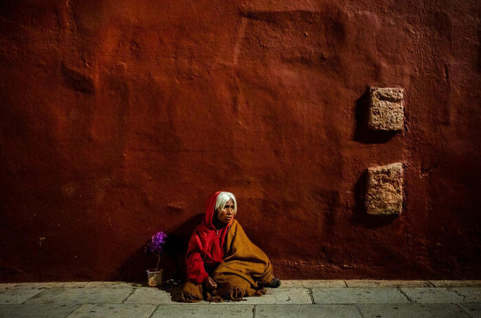 Elderly woman sitting against a textured wall in a candid street moment capturing the beauty of everyday life.