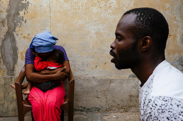 Mother embracing child seated on a chair while a man silently observes in an honest street moment capturing everyday life.