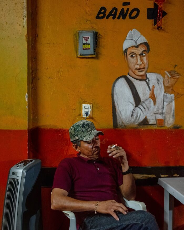 Man in camouflage hat smoking indoors beside wall painting, a street moment showing the beauty of everyday life.