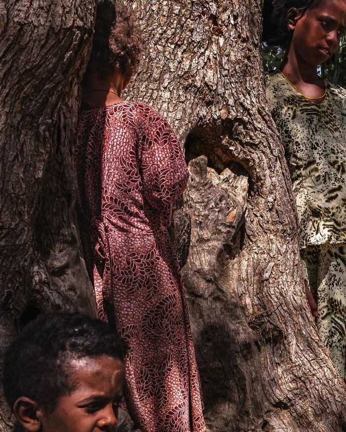 Three children in patterned clothing standing by a large tree, capturing honest street moments of everyday life.