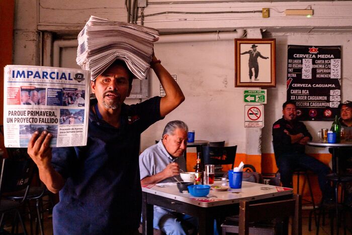 Man carrying stack of newspapers on his head in a busy indoor setting, capturing honest street moments of everyday life.