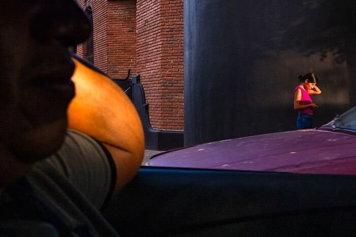 Man inside car in foreground and woman in pink top on street, capturing honest street moments that show everyday life beauty.