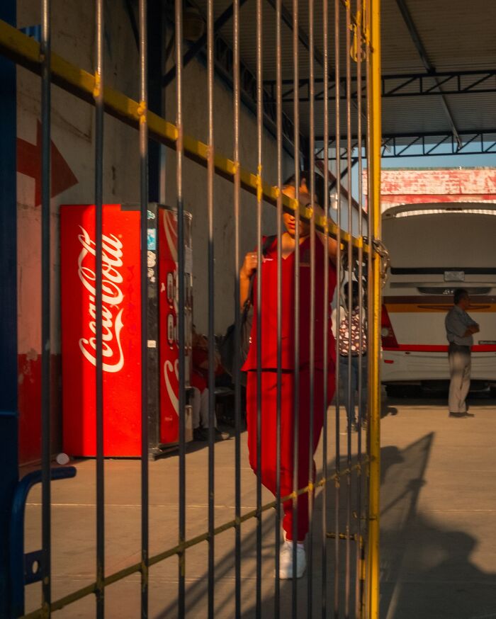 Person in red outfit walking behind yellow gate bars in a candid street moment showing the beauty of everyday life.