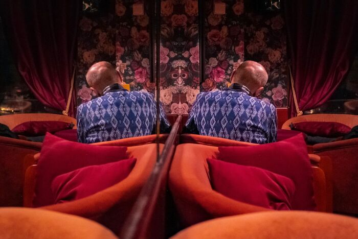 Elderly man in blue sweater seated by a floral wall and red cushions, capturing an honest street moment of everyday life.