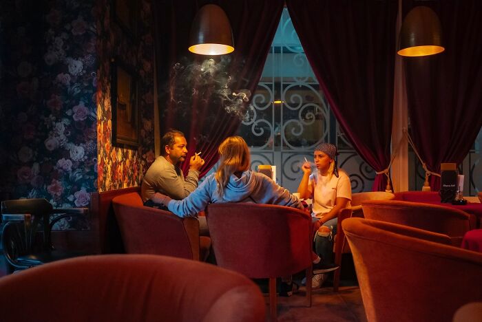 Three people sharing honest street moments inside a cozy room with warm lighting and vintage floral wallpaper.