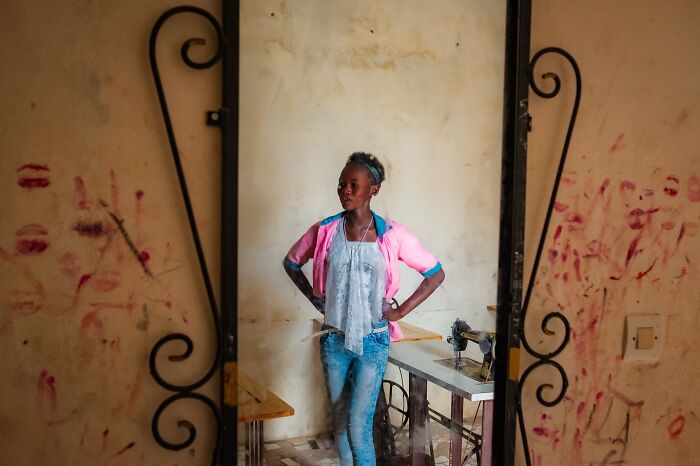 Young woman posing by a sewing machine in a rustic room, capturing an honest street moment of everyday life.