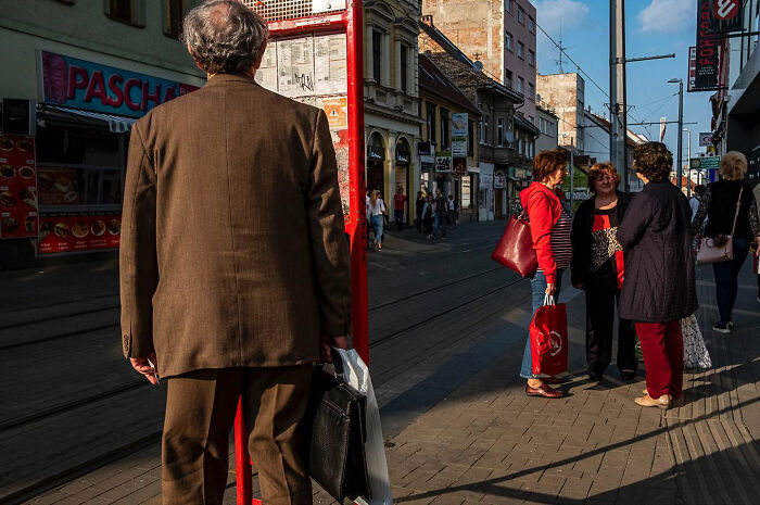 Man in brown suit waiting by tram stop while women chat on sunlit city street, capturing honest street moments of everyday life.
