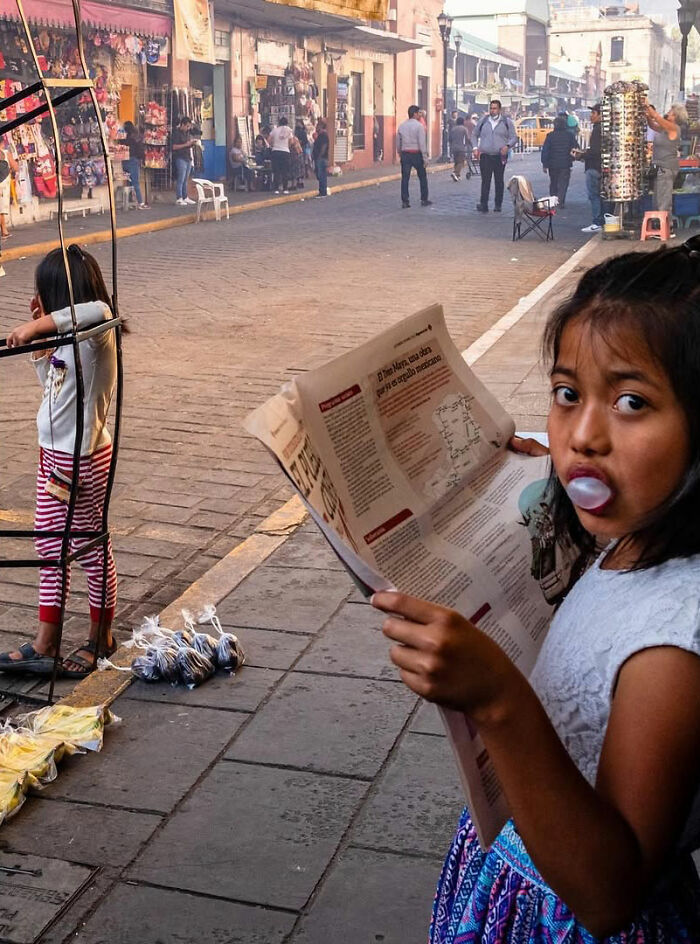 Young girl blowing bubblegum and reading a newspaper in an honest street moment capturing everyday life beauty.