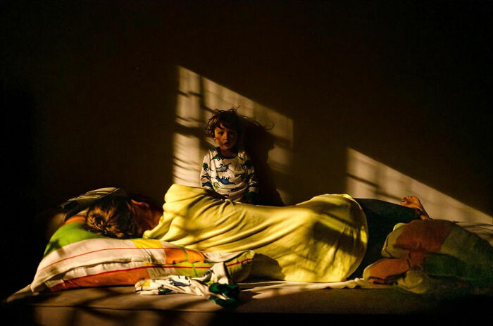 Child sitting on a bed next to a sleeping adult, warm light casting shadows, capturing honest street moments of everyday life.