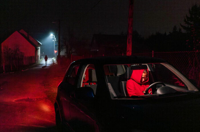 Person in a red hoodie sitting in a parked car at night on a dimly lit street, capturing honest street moments.