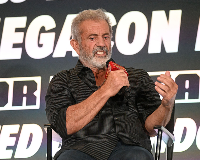 Hombre mayor con camisa negra hablando en micrófono durante evento sobre celebridades que arruinaron su reputación.