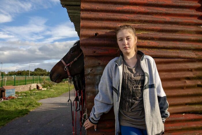 Irish Traveller kid leaning against a corrugated metal wall with a horse in a rural outdoor setting.