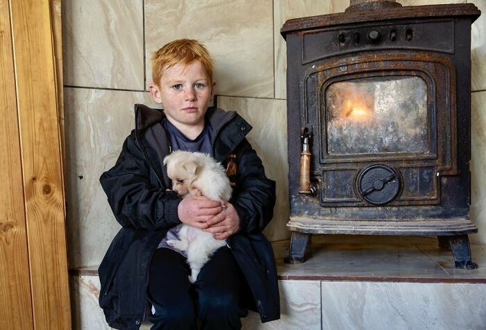 Irish traveller kid with red hair holding a puppy, sitting near a lit wood stove inside a home.