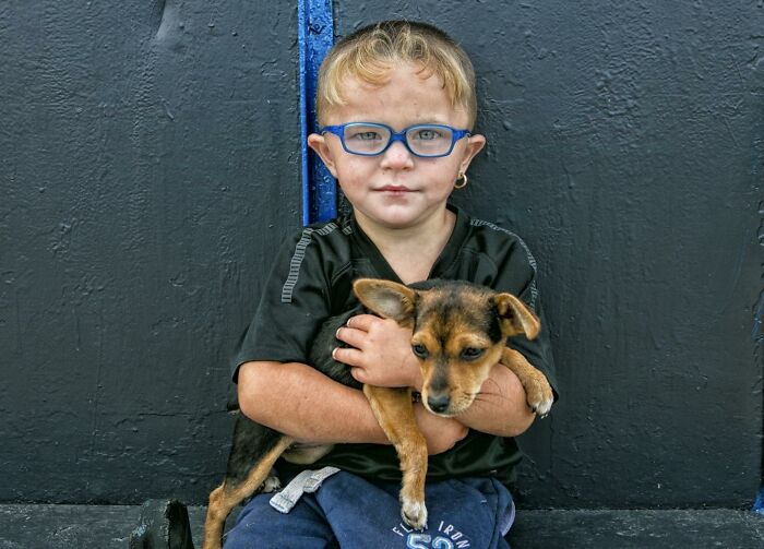 Irish Traveller kid with glasses holding a small brown dog, sitting against a dark textured wall.
