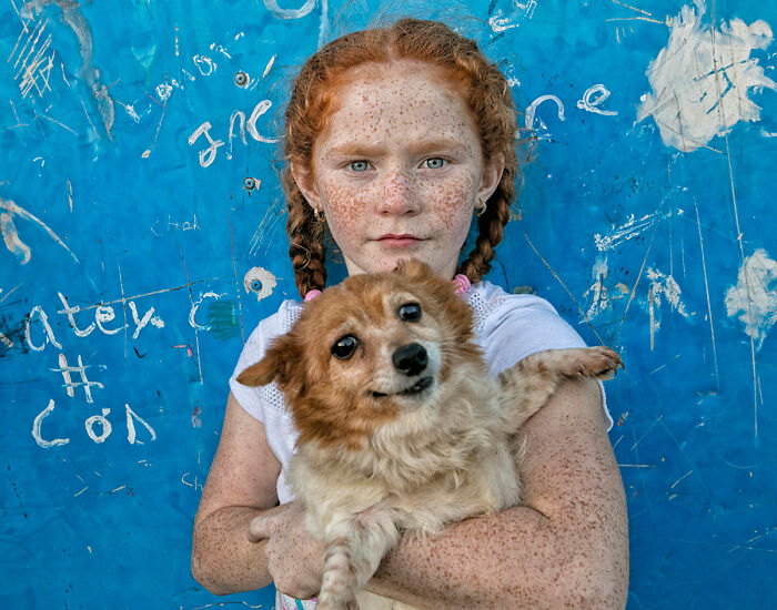 Irish Traveller child with red hair and freckles holding a small brown dog against a blue graffiti wall.