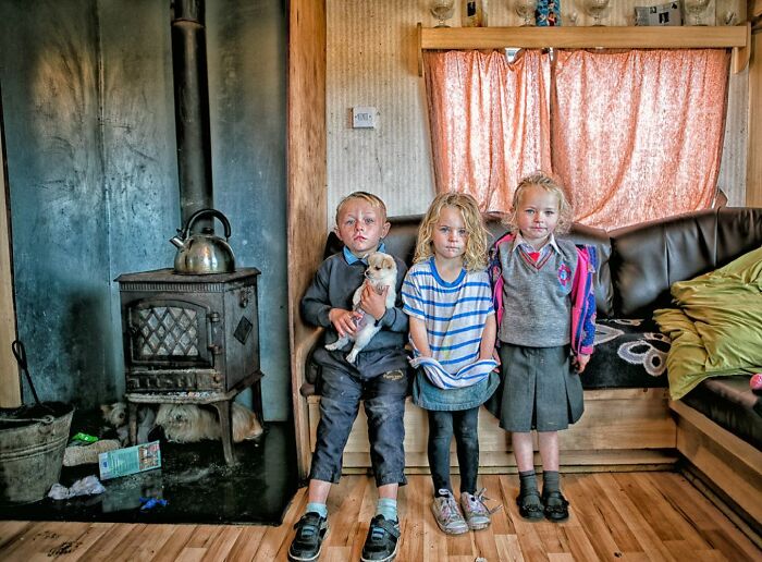 Three Irish Traveller kids indoors, one holding a puppy, sitting near a stove and wooden floor with cozy furnishings around.