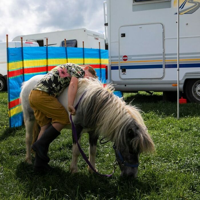 Irish Traveller kid hugging a small pony beside a campervan on green grass with a colorful windbreak nearby