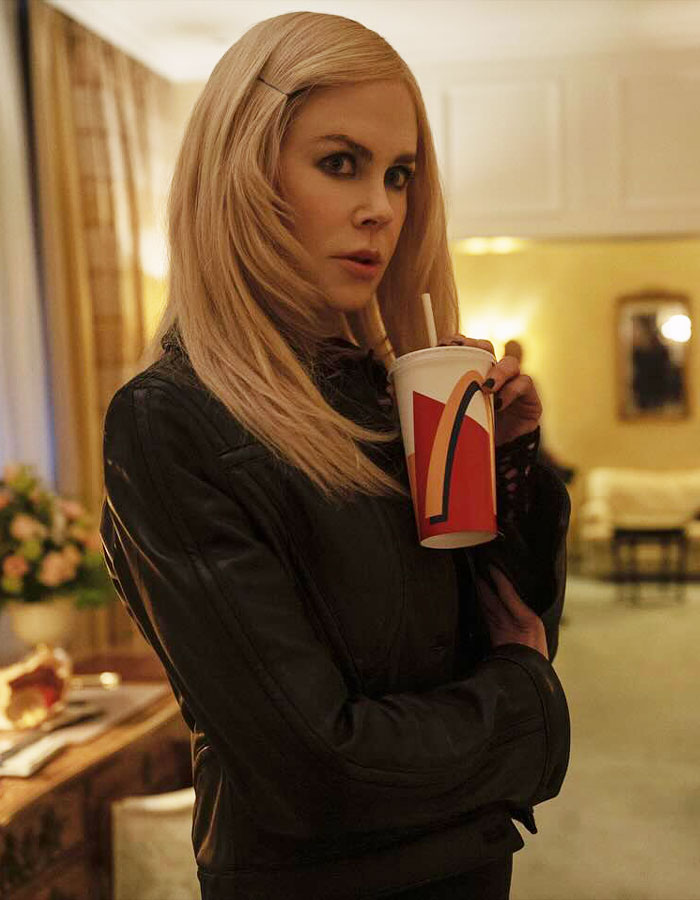 Nicole Kidman in a black leather jacket, holding a fast-food drink cup, with straight blonde hair indoors.