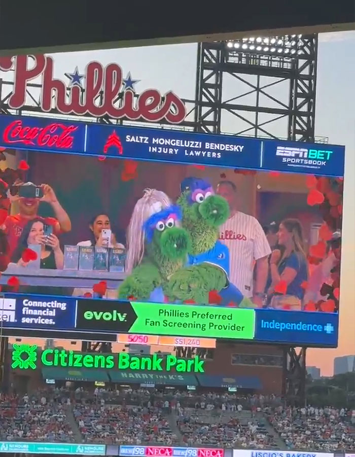 Baseball mascot in Phillies stadium humorously mocking CEO during kiss cam scandal on large digital screen.