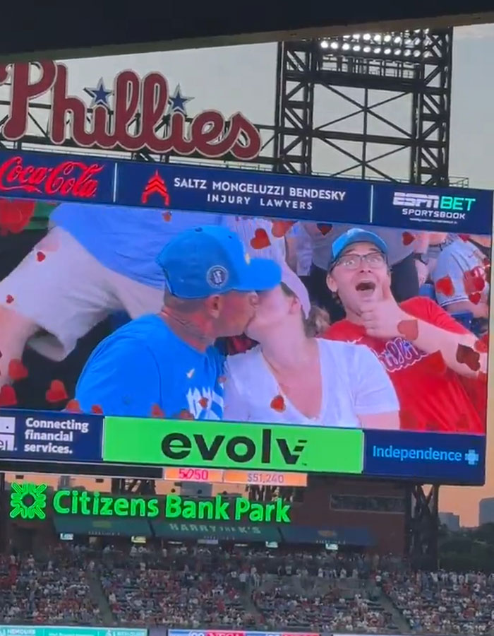 Baseball mascot humorously mocking disgraced CEO on kiss cam with fans kissing at Phillies game scoreboard.