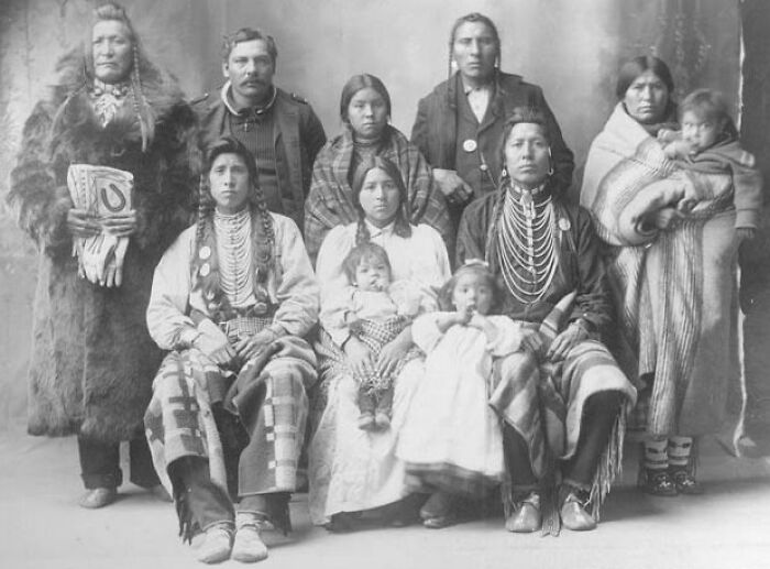 Vintage Native American family portrait with men, women, and children dressed in traditional clothing from the early 1900s.