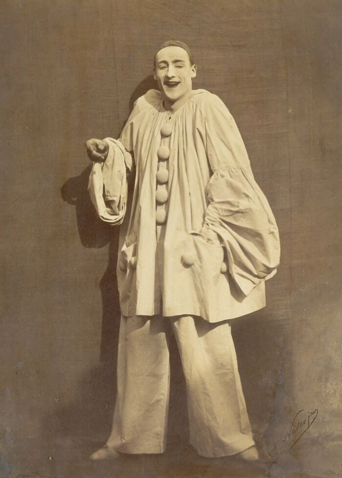 19th century photo of a person dressed as a clown with oversized buttons and wide pants in a sepia tone background.
