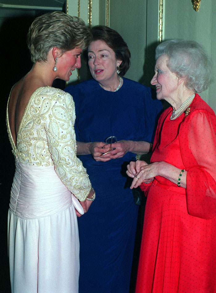 Three women in formal attire engaged in conversation at an elegant indoor event with ornate decor.