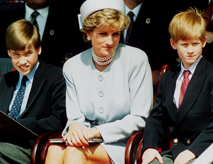 Princess Diana sitting between Prince William and Prince Harry at an outdoor event, all dressed formally in suits and a white outfit.