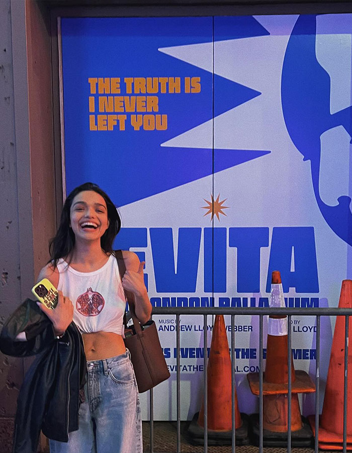 Rachel Zegler smiling and posing outside Evita theater with large blue and white promotional poster in background. Rachel Zegler smiling and posing outside Evita theater with large blue and white promotional poster in background.