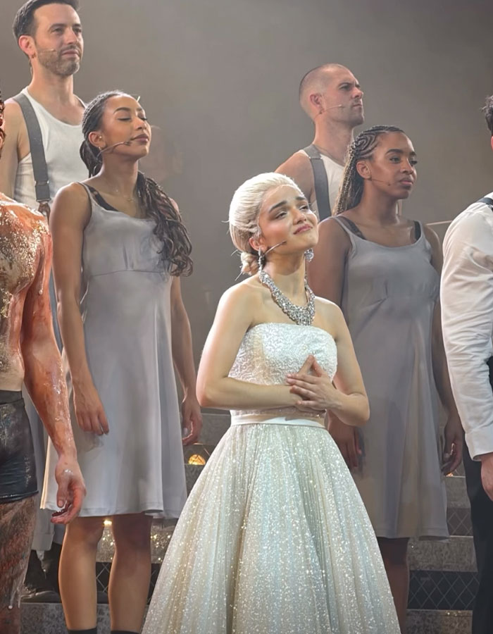 Understudy performing Evita on stage in a sparkling white dress, with cast members in gray costumes during a theater show. Understudy performing Evita on stage in a sparkling white dress, with cast members in gray costumes during a theater show.