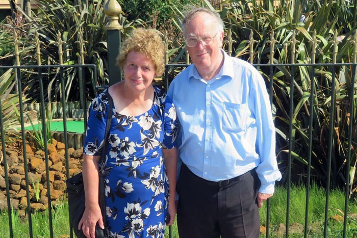 Older couple standing outdoors near plants and fence, related to documentary about woman living with deceased parents. - 4