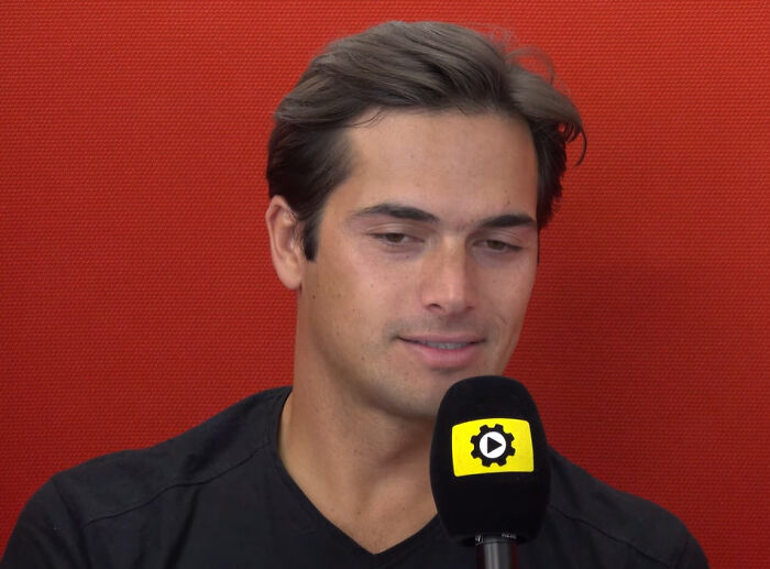 Man holding a microphone with a gear and play button logo, speaking against a red textured background about rumors.