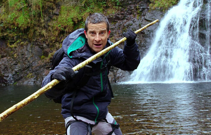 Man in outdoor gear holding a walking stick near a waterfall demonstrating survival tips in nature.