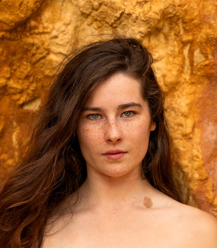 Gen Z woman with long hair and freckles standing against a textured orange background, symbolizing natural beauty and confidence.