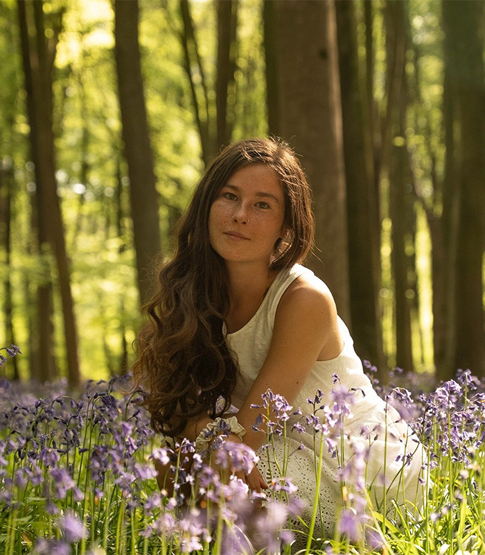 Gen Z woman with long hair sitting in a forest surrounded by purple flowers, embracing natural beauty and body hair.