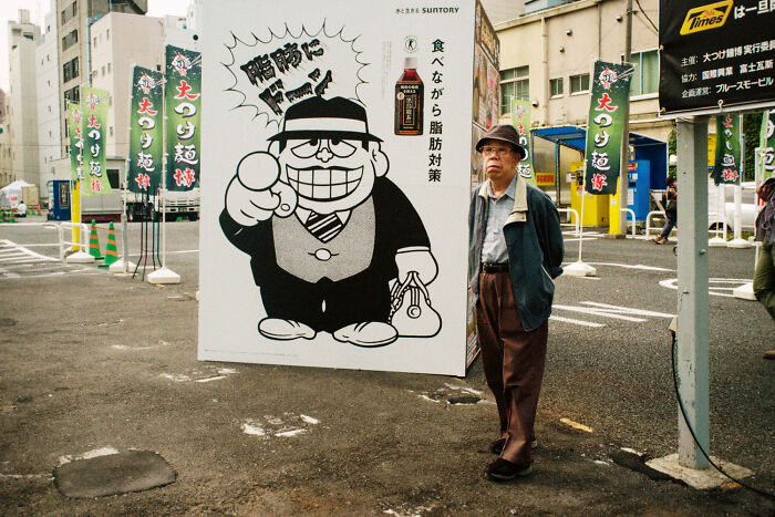 Elderly man walking near a street advertisement in a Japanese urban setting showing daily life in Japan by Shin Noguchi.