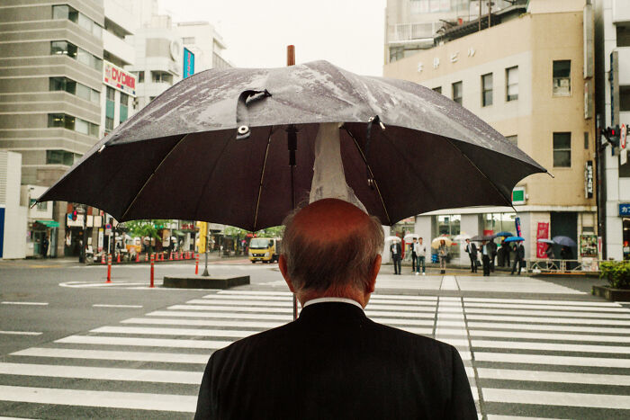 Elderly man with umbrella crossing a busy urban street, capturing daily life in Japan by photographer Shin Noguchi.
