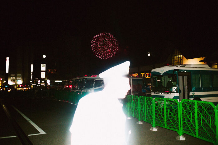 Night scene of daily life in Japan showing a bright silhouette of a police officer with fireworks in the background.
