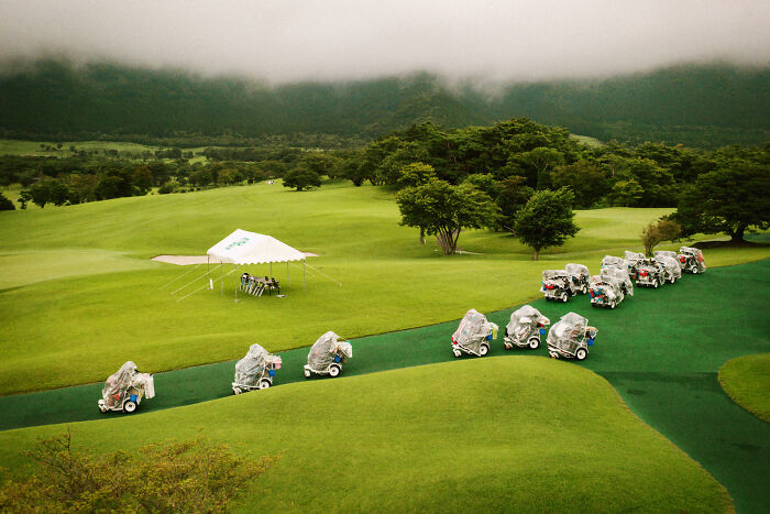 Golf carts covered in rain protection lined up on a green course, depicting daily life in Japan by Shin Noguchi.