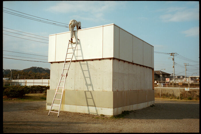 Person climbing ladder to rooftop of a concrete building, depicting daily life in Japan by photographer Shin Noguchi.
